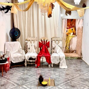 May include: An interior shot of a room decorated for a ceremony. Three chairs are adorned with white and red bows. A chandelier hangs from a gold draped ceiling. A small table holds a vase and bowl. The floor is tiled.