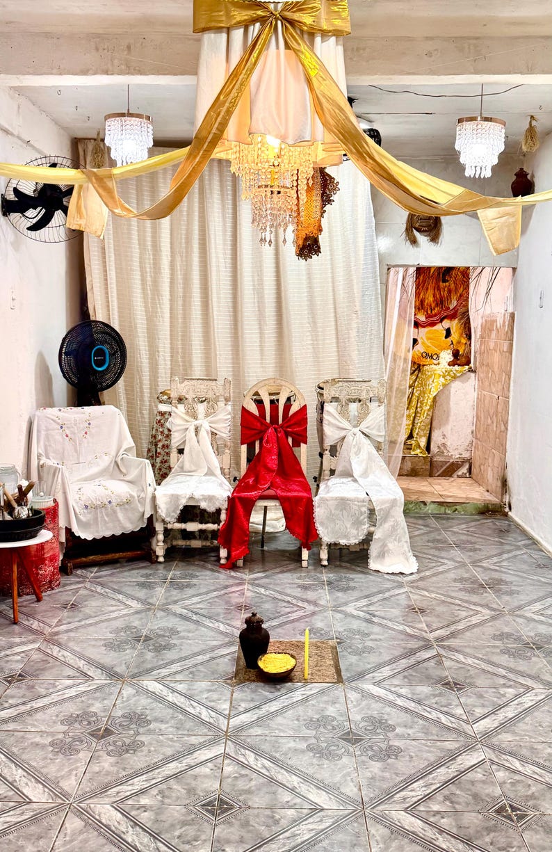 May include: An interior shot of a room decorated for a ceremony. Three chairs are adorned with white and red bows. A chandelier hangs from a gold draped ceiling. A small table holds a vase and bowl. The floor is tiled.