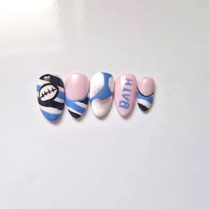 May include: A set of five almond-shaped press-on nails featuring a light blue, black, and white colour palette. The designs include horizontal stripes, a football graphic, and the word BATH written in blue capital letters on a light pink base.