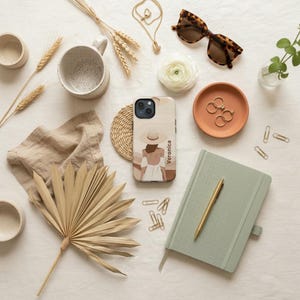 May include: A smartphone case featuring an illustration of a person wearing a sun hat and white dress with the name Veronica printed vertically. The case is surrounded by a sage green notebook, a gold pen, tortoise shell sunglasses, and dried botanicals.