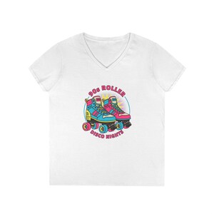 Puede incluir: Camiseta blanca con cuello en V con un gráfico colorido de patines retro. El diseño incluye el texto "90s Roller Disco Nights" en un diseño circular. Los patines son en tonos de azul, rosa y amarillo.