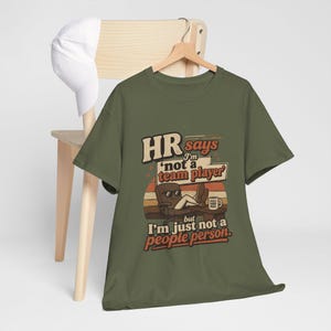 May include: An olive green short-sleeve t-shirt featuring a graphic print with the text "HR says I'm 'not a team player' but I'm just not a people person." The design includes a cartoon character in an office chair with feet on a desk.