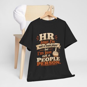 May include: A black short-sleeved t-shirt featuring the text HR says I'm not a team player but I'm just not a PEOPLE PERSON with a graphic of a hand showing the middle finger. The shirt is displayed on a wooden hanger with a white baseball cap.