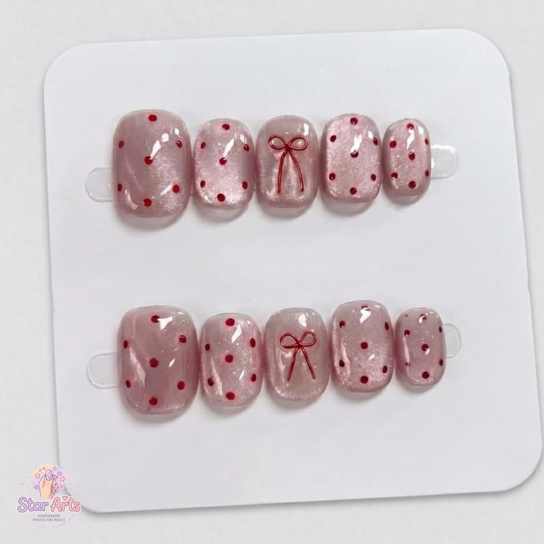 May include: A set of press-on nails with a light pink base, adorned with small red polka dots and a red bow design on some nails. The nails are oval-shaped and come in various sizes, presented on a white card.