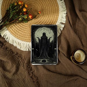 May include: A black and white illustration of a robed figure with outstretched arms, set against a full moon and a dark forest. The image is on a rectangular object, possibly a phone case or a book cover. A straw hat and a candle are also in the image.