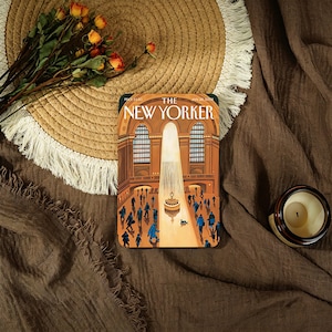 Puede incluir: Un objeto rectangular decorativo con una ilustración de la portada de la revista New Yorker que representa una gran sala con gente. El objeto está sobre una superficie marrón, junto a un sombrero de paja, flores y una vela.
