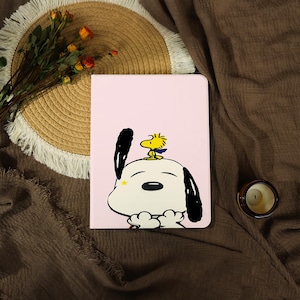 Op de afbeelding: Een lichtroze tablethoes met een grafische illustratie van een witte hond met zwarte oren en een kleine gele vogel op zijn kop. De hond heeft een klein geel sterretje op zijn wang en houdt zijn pootjes bij elkaar voor zijn kin.