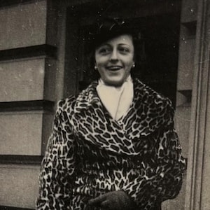 May include: A woman wearing a black and white leopard print coat with a white scarf and a black hat. She is standing in front of a building with a black iron fence.