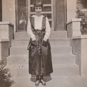 Może przedstawiać: Czarno-biała fotografia osoby pozującej przed drzwiami. Osoba ma na sobie długą, ciemną sukienkę z kieszeniami, białą bluzkę i ciemne buty. Obraz ma wygląd vintage, z lekko wyblakłym wyglądem.