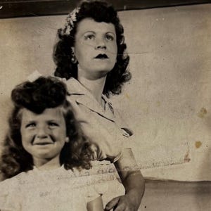 May include: A black and white photo of a mother and daughter. The mother is wearing a light-colored dress and has her hair styled in a bouffant. The daughter is wearing a white dress and has her hair in a ponytail. The mother is smiling and looking at the camera, while the daughter is looking at the camera and smiling.