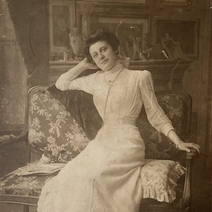 May include: A woman in a white dress sits on a floral patterned couch with her arm resting on the back. She is wearing a pearl necklace and has her hair pulled back.