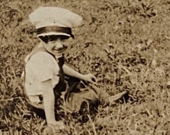 May include: A young boy wearing a white cap sits in a field of tall grass. He is smiling and looking at the camera.