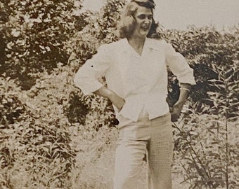 May include: A woman in a white button-down shirt and white pants stands in a field of tall grass. She is smiling and looking to the right. There are trees in the background.