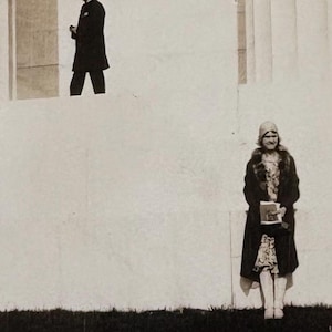 May include: A black and white photograph of a man in a uniform and a woman in a fur-trimmed coat standing in front of a building.