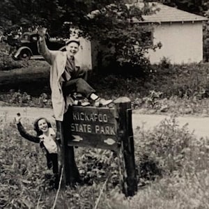 May include: A black and white photo of two people standing in front of a sign that reads "Kickapoo State Park". The people are smiling and waving. The sign is made of wood and has a brown background.
