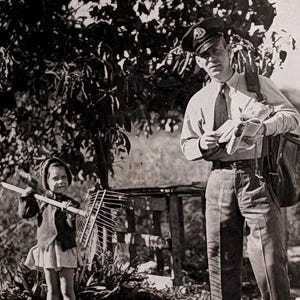 Pode incluir: Fotografia vintage a preto e branco de um carteiro de uniforme, segurando cartas e uma bolsa, num jardim. Uma criança com um ancinho está por perto. A cena é ao ar livre com uma árvore e folhagem.