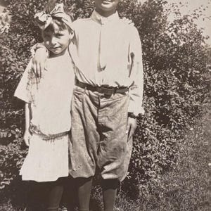 May include: Vintage black and white photograph of two children posing outdoors. One child wears a white shirt, knickerbockers, and a hat. The other child wears a white dress and a bow in their hair. They are standing in front of a bush.