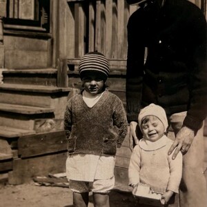 May include: A vintage black and white photograph showing a person wearing a dark cardigan and a fedora hat standing behind two children. One child wears a striped beanie, a V-neck sweater, and knee-high socks. The other child wears a light-colored cap.
