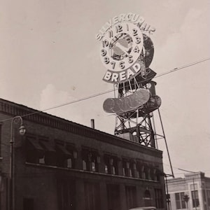 May include: Vintage black and white photograph of a building with a large clock-shaped sign that reads "Silvercup Bread." Classic cars are parked along the street in front of the building. The sky is overcast.