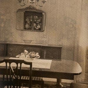 May include: Sepia-toned dining room scene featuring a dark wooden table and chairs. A chandelier hangs above, and a mirror is mounted on the wall. A buffet and a floral arrangement complete the setting, creating a vintage aesthetic.