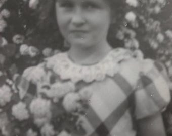 May include: A young girl wearing a white lace collar and a checkered dress, with a bouquet of flowers in her hands. She is standing in front of a background of white flowers.