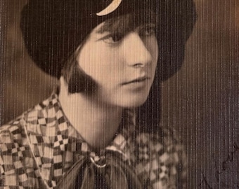 May include: A vintage sepia-toned photograph featuring a person wearing a dark, rounded beret-style hat adorned with a small, light-colored crescent moon emblem. The individual wears a collared shirt with a geometric checkered pattern and a necktie.