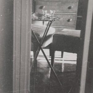 Puede incluir: Una fotografía en blanco y negro de una habitación con una mesa pequeña, dos sillas y una cómoda. La mesa tiene una bandeja decorativa con dos vasos. La cómoda tiene varios cajones con pomos redondos. La escena se refleja en un espejo.