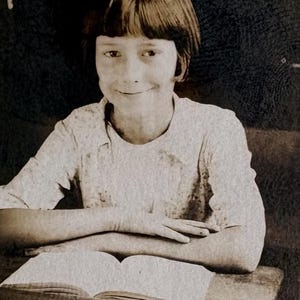 May include: A sepia-toned photograph of a young person seated at a desk, smiling at the camera. The person has short, dark hair and is wearing a light-colored shirt. An open book rests on the desk in front of them, with their arms crossed over it.