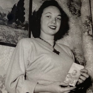 Puede incluir: Una fotografía en blanco y negro de una mujer que lleva un suéter de color claro y un collar. Ella sostiene una tarjeta pequeña en su mano derecha. El fondo es un papel tapiz estampado.