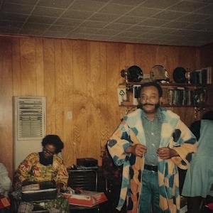 Puede incluir: Dos personas están en una habitación con paneles de madera. La persona de la derecha lleva una bata de color azul y blanco con estampado. La persona de la izquierda lleva una camisa de color amarillo y marrón con estampado.