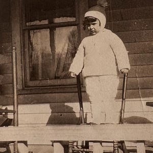May include: A vintage sepia-toned photograph of a child wearing a knit sweater, matching trousers, and a pom-pom beanie. The child stands on a wooden porch, holding onto the back of a wooden chair, with a window visible in the background.