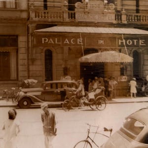 May include: A sepia-toned vintage photograph depicting the exterior of the Palace Hotel with the text Palace Hotel visible on the building facade. The street scene features a classic automobile, a three-wheeled cycle, bicycles, and pedestrians.