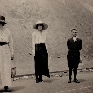 Peut inclure: Photographie en noir et blanc de quatre personnes sur une route. Deux femmes portent des chapeaux et de longues robes. Un garçon en costume et une fille en robe et chapeau se tiennent à proximité. En arrière-plan, une colline.