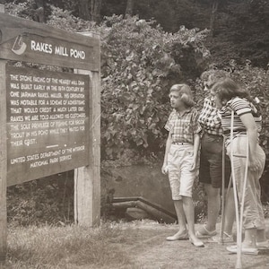 May include: A black and white photo of a sign that reads "Rakes Mill Pond". The sign explains the history of the mill and pond. Three people are standing in front of the sign, looking at it. The sign is made of wood and has a metal frame.