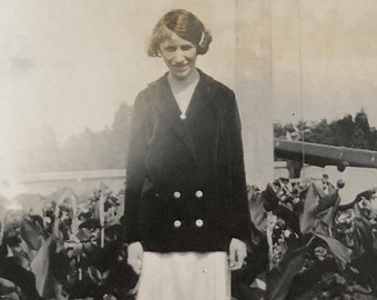 May include: A black and white photo of a woman wearing a black jacket with double breasted buttons and a white skirt. She is standing in front of a garden.