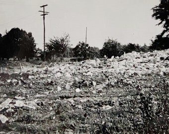 May include: A black and white photo of a pile of rubble and debris in a field. There are trees in the background and a utility pole with wires.