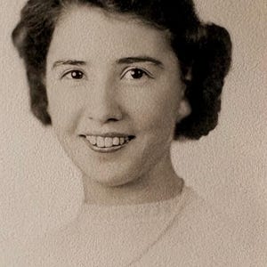 May include: Black and white portrait of a woman with dark, wavy hair and a warm smile. She is wearing a light-colored top. The image has a vintage feel.
