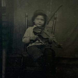Può includere: Fotografia in bianco e nero di un bambino che suona il violino. Il bambino, con un cappello e abiti chiari, è seduto su una sedia di legno. Il violino è tenuto contro il mento del bambino, con l'arco sulle corde.