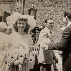 Può includere: Una sposa in abito bianco e un grande cappello bianco tiene un bouquet di fiori bianchi. È circondata da ospiti durante una cerimonia di matrimonio.