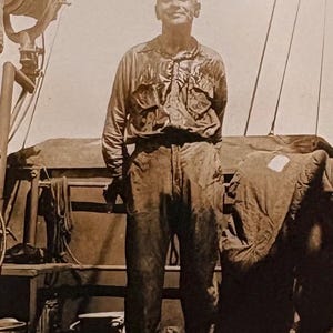 May include: A sepia-toned photograph of a man standing on a ship deck. He wears a long-sleeved shirt and pants, both appearing wet. The man is smiling, with his hands behind his back. The background includes ship equipment and rigging.