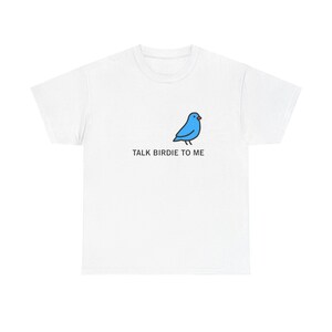 Könnte beinhalten: Weißes T-Shirt mit einem blauen Cartoon-Vogel und dem Text "TALK BIRDIE TO ME". Der Vogel ist einfach gezeichnet, und der Text ist in einer klaren Schriftart. Das T-Shirt besteht aus einem weichen Material, das sich für den lässigen Gebrauch eignet.