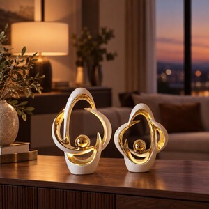 Peut inclure: Deux sculptures abstraites présentant un extérieur blanc lisse et un intérieur doré réfléchissant. Chaque pièce a une forme organique incurvée avec une sphère dorée centrale suspendue dans le cadre ouvert, créant un ensemble décoratif équilibré.