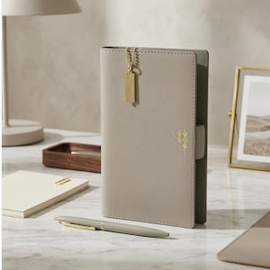 Peut inclure: Un journal en cuir gris clair avec une chaîne et une étiquette dorées attachées. Un stylo assorti avec des accents dorés repose sur la surface. Le journal a un motif géométrique et un fermoir. Un bloc-notes et un cadre photo sont également visibles.