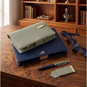 May include: Two leather-bound journals, one olive green and one navy blue, are stacked on a wooden surface. A matching pen and a green and gold money clip are next to the journals. The journals have gold accents and a pen holder.