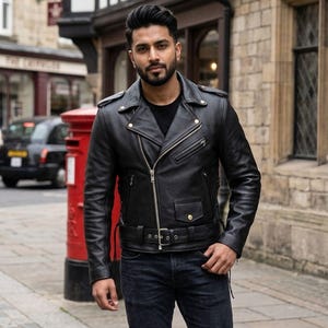 Op de afbeelding: Een zwarte leren jas in klassieke bikerstijl. De jas heeft een reverskraag, een asymmetrische rits en een ceintuur in de taille. Het model draagt een donkerblauwe jeans en een zwart T-shirt.