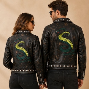Op de afbeelding: Twee zwarte leren jacks met een verweerde uitstraling en een grote geborduurde slang in groen- en geeltinten op de rug. De jacks zijn voorzien van zilverkleurige metalen studs langs de kraag en zoom, lange mouwen en zilveren ritsen.