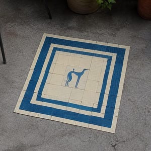 May include: A square rug with a blue and white design. The rug features a blue border and a central image of a greyhound dog. The rug is made of woven material and is placed on a concrete surface.