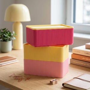 Könnte beinhalten: Ein Stapel aus drei rechteckigen Aufbewahrungsboxen mit vertikaler Riffelstruktur. Die oberste Box ist magentafarben mit einem gelben Deckel, die mittlere Box ist gelb und die untere Box ist hellrosa.
