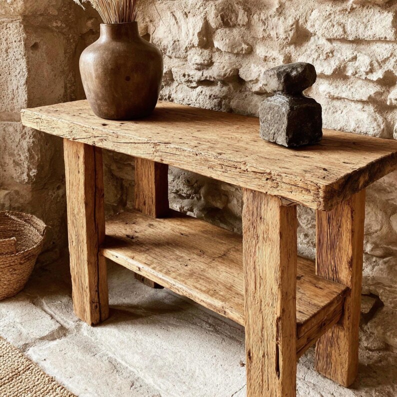 Op de afbeelding: Een rustieke houten console tafel met een onderste plank. De tafel is gemaakt van gerecycled hout en toont natuurlijke houtnerf en textuur. Een bruine vaas en een donkergrijze stenen sculptuur staan op het tafelblad. De tafel is ongeveer 120 cm lang.