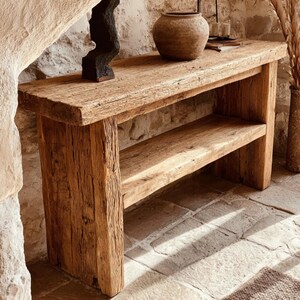 Op de afbeelding: Een rustieke houten console met een onderste plank. Het hout toont natuurlijke nerven en textuur. Een donkere sculptuur en een keramische vaas staan op het tafelblad. De console staat tegen een stenen muur. Ongeveer 150 cm lang.